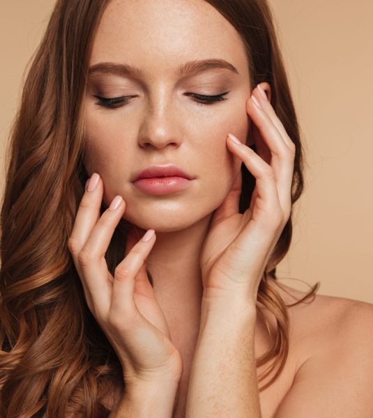 Close up Beauty portrait of sensual ginger woman with long hair posing with arms near the face over cream background