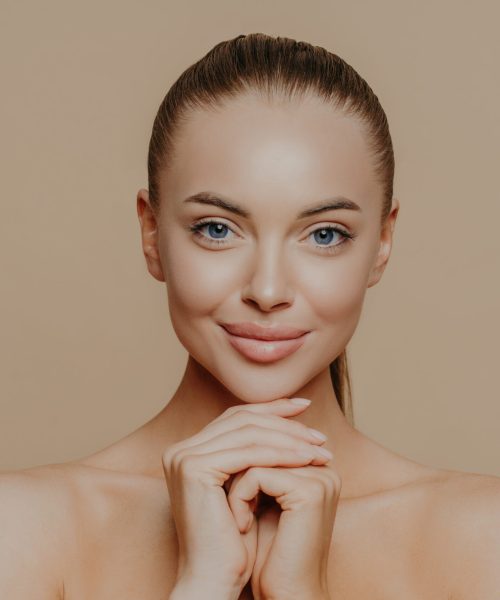 Young pretty woman with beauty skin and combed hair, keeps hands under chin, has healthy complexion after face purification, soft well cared body, isolated over studio wall. Facial treatment